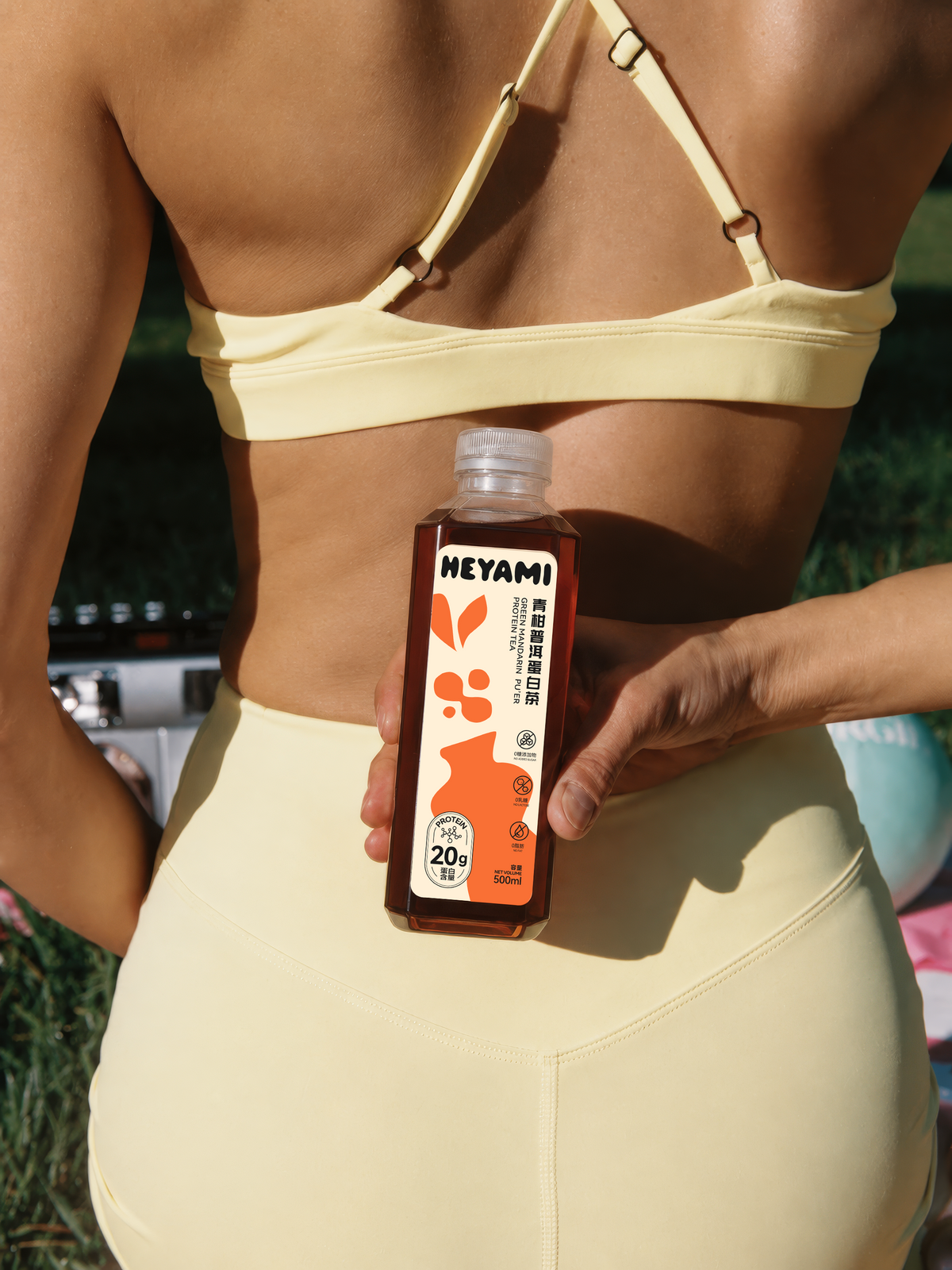 A woman outdoors wearing a pale yellow bikini and holding a bottle of Heyami protein tea behind her back. The bottle has an orange and white label with illustrations of Green Mandarin Puer.